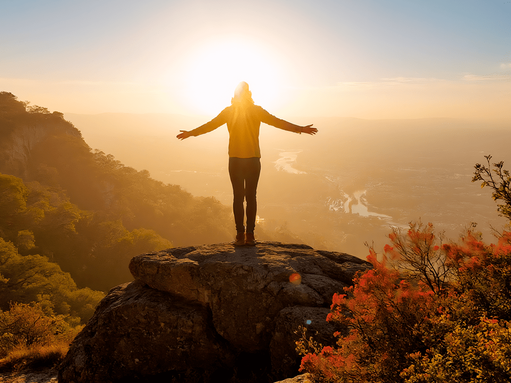 Mentalidade de abundância representada por pessoa com braços abertos no topo de uma montanha ao nascer do sol, simbolizando gratidão, crescimento e positividade.